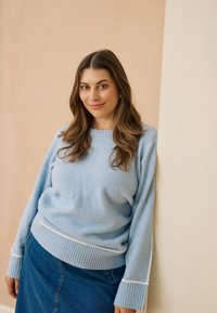 Pull bleu clair texturé avec poignets et ourlet côtelés, associé à une jupe en denim avec une fente frontale et des détails de couture ton sur ton.