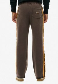 Brune sweatpants lavet af blødt materiale, med orange side-striber, en baglomme og en afslappet pasform. Gummisål fodtøj synligt.