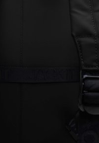 Zaino nero con texture liscia, dotato di una prominente cintura piana etichettata "JACKIT" e di una fibbia resistente con una finitura a contrasto.