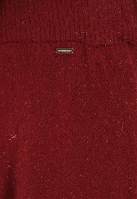 Pulover tricotat de culoare burgundă, cu o textură moale, o strălucire subtilă și un etichetă mică cu logo auriu. Materialul prezintă fire fine și un guler ribat.