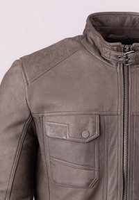 Brown leather jacket with a stand-up collar, zip closure, and two chest pockets. Features textured shoulder panels and metal accents.