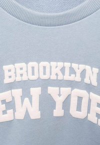 Světle modrá mikina s bílým vyvýšeným nápisem "BROOKLYN NEW YORK". Hladký materiál a žebrovaný výstřih.