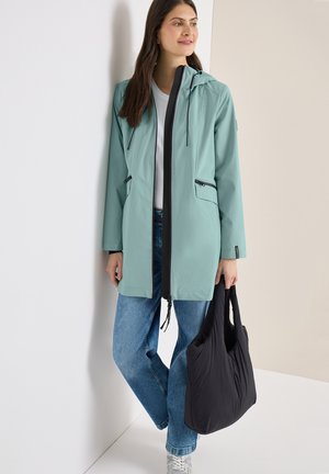 Femme portant une veste à capuche vert clair, chemise blanche, jean bleu, baskets blanches, tenant un grand sac fourre-tout noir, appuyée contre un mur.
