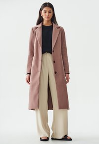 Cappotto lungo di un leggero colore rosa chiaro realizzato in un tessuto testurizzato, con colletto a risvolto, bottoni monopetto e una silhouette dritta, abbinato a pantaloni beige.