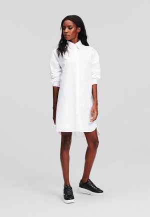 Weißes Hemdkleid aus glattem Stoff, mit langen Ärmeln, Knopfverschluss vorne und seitlichen Schlitzen. Kombiniert mit schwarzen Turnschuhen.