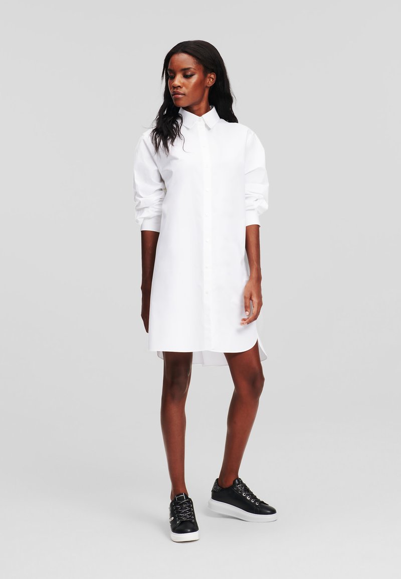 Abito camicia bianco con colletto realizzato in tessuto morbido, dotato di maniche lunghe, chiusura con bottoni e spacco laterale. Abbinato a sneakers nere.