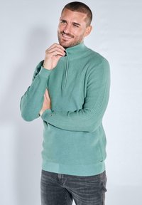 Hellgrüner, strukturierter Strickpullover mit hohem Kragen und halbem Reißverschluss. Gerippte Bündchen und Saum, mit kleinem Logodetail an der Seite.