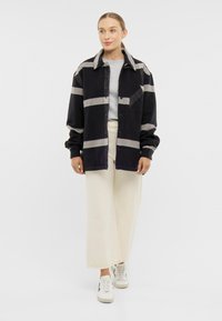 Černá a šedá pruhovaná košile na knoflíky s límcem, s kapsou na hrudi, spojená s béžovými širokými kalhotami a bílými teniskami.