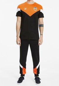 Hombre vistiendo un conjunto deportivo de Valencia CF de Puma en negro y naranja, con el logo en el pecho, joggers negros con acentos en naranja y blanco, y zapatillas negras.