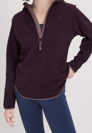 Kobieta nosząca ciemnofioletową bluzę polarową z zamkiem do ćwiartki oraz granatowe legginsy, stojąca z jedną ręką na biodrze, drugą rozluźnioną.