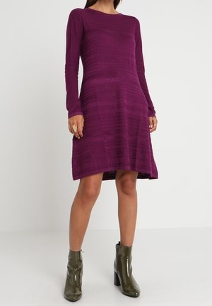 Mujer con un vestido morado de manga larga hasta la rodilla y botines brillantes verde oliva, de pie frente a un fondo blanco liso.