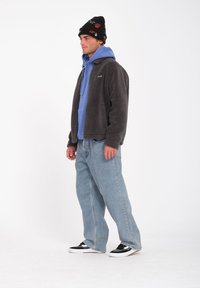 Conjunto masculino que incluye una chaqueta gris de felpa y una sudadera con capucha azul, combinada con unos vaqueros anchos de color azul claro y zapatillas negras. Gorro negro con acentos coloridos.