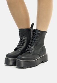 Botas de tobillo de cuero negro con una suela de plataforma alta, parte frontal con cordones, cremallera lateral y detalle de costura en contraste. Suela de goma texturizada.