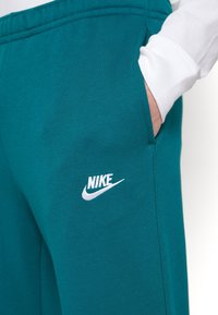 Tyrkysové tepláky vyrobené z měkké látky, se bílým logem Nike na levém stehně a bočním kapsou. Elastický pas a uvolněný střih.