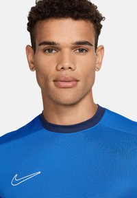 Blauwe T-shirt met korte mouwen en een ronde halslijn, met donkerder blauwe accenten. Nike-logo in het wit aan de linkerkant. Glad, aansluitend textuur.