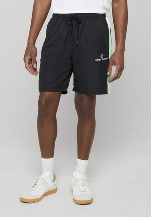 Pantalones cortos deportivos negros para hombre con paneles laterales en color menta, logotipo blanco de "Sergio Tacchini" en la pierna derecha, usados con calcetines blancos y zapatillas blancas.
