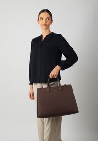 Brown leather handbag with smooth texture, rounded handles, and gold hardware. Worn by a person in a black blouse and beige pants.