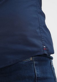 Detail blízkého záběru na lem námořnické modré košile přes tmavě modré džíny, ukazující malé červené, bílé a modré prošití u bočního švu.