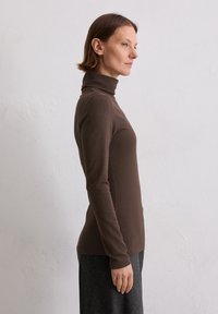 Brauner Rollkragenpullover in taillierter Form mit langen Ärmeln und einer weichen Textur, kombiniert mit einem dunkelgrauen Rock. Seitenansicht vor einem hellen Hintergrund.