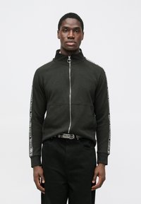 Czarna kurtka na zamek z wysokim kołnierzem, srebrnym suwakiem i taśmą z logo na rękawach. Noszona z czarnymi spodniami i wzorzystym paskiem.