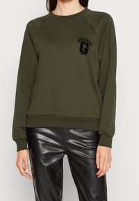 Persona che indossa una felpa verde oliva con logo nero "Athletic Dept" e pantaloni in pelle nera lucida, in piedi contro uno sfondo bianco.