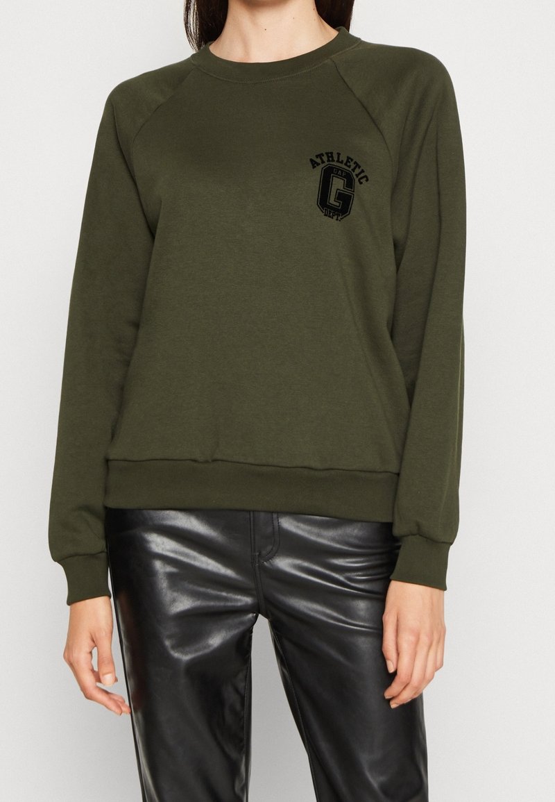 Persona che indossa una felpa verde oliva con logo nero "Athletic Dept" e pantaloni in pelle nera lucida, in piedi contro uno sfondo bianco.