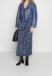 Blå lädercroppad bikerjacka över en svart klänning med blommönster och blå blommor, kompletterad med svarta glänsande knähöga stövlar och en svart väska.