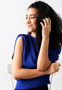 Kungligt blå kortärmad topp med draperad tygdetalj, mjuk textur och rund halsringning. Modell med lockigt hår poserar och vilar en hand på sin axel.