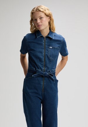 Blondynka ubrana w krótkirękawowy ciemnoniebieski kombinezon dżinsowy z przednim zamkiem i paskiem, stojąca na jednolitym jasnym tle.