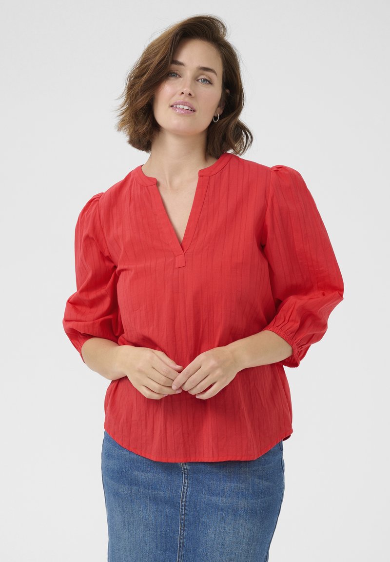 Blusa rossa con texture, scollatura a V e maniche a palloncino, abbinata a una gonna in denim blu. La camicia presenta sottili righe verticali.