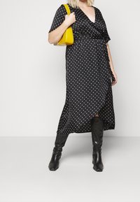 Schwarzes Wickelkleid mit Punkte, kurzen Ärmeln, V-Ausschnitt und seitlichem Schlitz, kombiniert mit einer gelben Handtasche und kniehohen schwarzen Stiefeln.