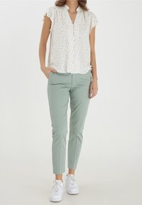 Weißes Hemd mit Blumenmuster und kurzen Rüschenärmeln, kombiniert mit schmal zulaufenden mintgrünen Hosen und weißen Turnschuhen.