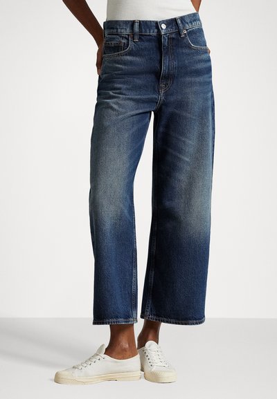 Πλατύ παντελόνι τζιν σε μπλε χρώμα με ήπιες φθορές, μπροστινές τσέπες και κλασικό κουμπί κλεισίματος. Συνδυασμένο με λευκά καμβά sneakers.