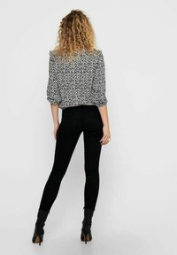 Nainen, jolla on kiharat hiukset, pukeutuneena musta-valkoiseen kukkakuosisiseen paitaan, mustiin tiukkoihin farkkuihin ja mustiin korkokenkiin, seisoo selkä kameraan päin yksinkertaisella taustalla.