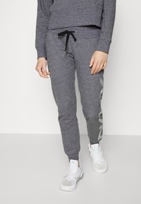 Šedé tepláky s zúženým střihem, s černým stahovacím šňůrkem a velkým světle šedým logem na levé straně. Hladká textura tkaniny.