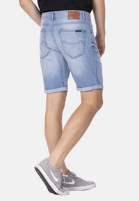 Shorts en denim bleu clair avec des revers, poches arrière et une étiquette en cuir marron sur la taille. Portés avec des baskets grises.