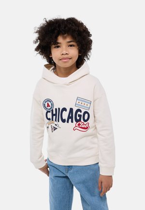 Jeune enfant aux cheveux bouclés portant un sweat à capuche blanc avec le texte « CHICAGO Club » et des écussons, ainsi qu'un jean bleu, debout devant un fond uni.
