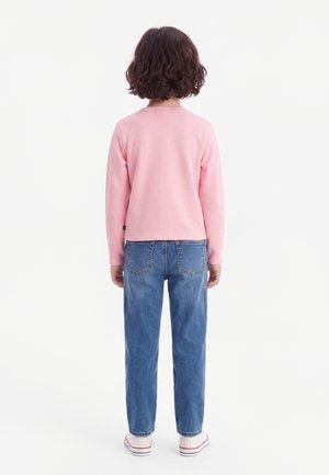 Enfant aux cheveux bouclés portant un t-shirt rose à manches longues, un jean bleu et des baskets blanches, debout de dos sur un fond blanc.