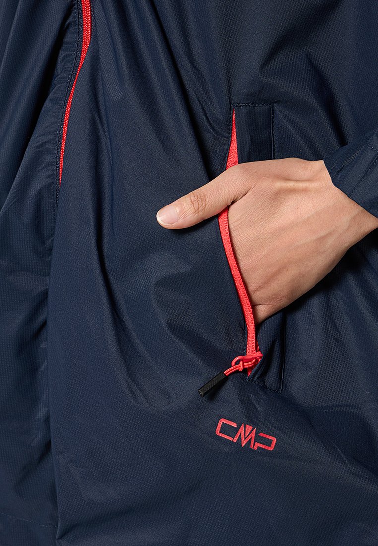 Mano dentro della tasca con zip di un giubbotto blu navy con rifiniture rosse e il logo CMP.