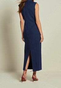 Robe sans manches bleu marine avec une fente haute, design ajusté, texture lisse et fermeture éclair au dos. Associée à des sandales à talons aiguilles rouges à brides.