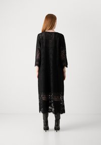 Black lace maxi dress with three-quarter sleeves, intricate patterns, and a scalloped hem. Worn with black heeled boots.