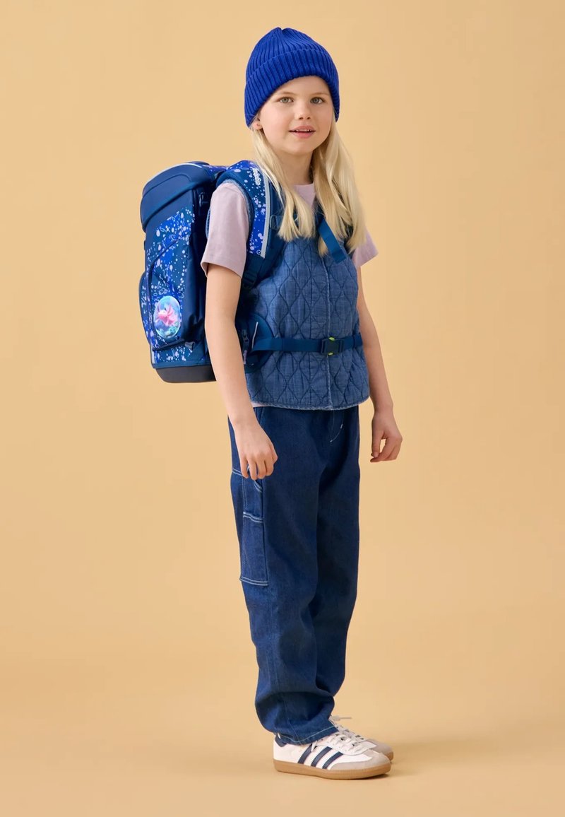 Gilet matelassé bleu sur une chemise violet clair, associé à un jean bleu large et des baskets blanches. Un sac à dos bleu à motifs complète la tenue.