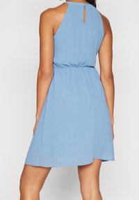 Vrouw die een mouwloze lichtblauwe jurk draagt met een geplooide taille en een sleutelgatdetail aan de achterkant, staand tegen een effen achtergrond.