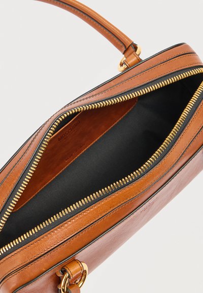 Καφέ δερμάτινη τσάντα με χρυσλό φερμουάρ που είναι μερικώς ανοιχτό, δείχνοντας μαύρο υφασμάτινο εσωτερικό και λογότυπο της μάρκας χαραγμένο στην εσωτερική πλευρά.