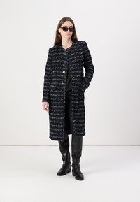 Svart och vit tweed lång kappa med ett strukturerat mönster, med rak skärning, utan knäppningar och sidofickor, bärs över svarta byxor och stövlar.