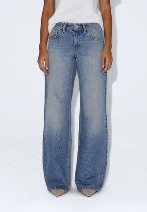 Woman wearing light blue wide-leg jeans with a high waist and beige pointed shoes, standing against a plain white background.