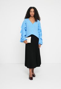 Blouse bleu clair avec des manches voluminieuses à volants, associée à une jupe plissée noire. Une pochette crème et des chaussures rouge foncé complètent le look.
