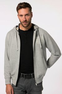 Sudadera gris con cremallera y cordón, presenta un forro interior a cuadros. Combinada con una camiseta henley negra y jeans grises.