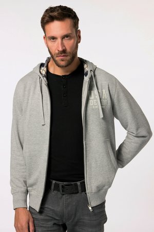 Felpa grigia con zip e cordino, presenta una fodera interna a scacchi. Abbinata a una maglietta henley nera e jeans grigi.