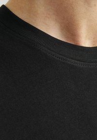 Črna bombažna majica s kratkimi rokavi, posnetek v bližini, ki prikazuje rebrast okrogli izrez in gladko teksturo s urejeno šivano podrobnostjo pri ovratniku.